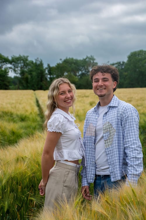 Un couple debout au milieu d’un champ de blé vert, souriant à l’objectif, lisière d’arbres et ciel nuageux derrière eux.